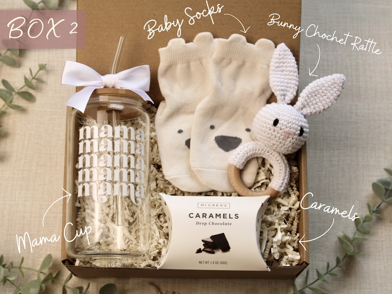 May include: A gift box filled with baby items, including a white crocheted bunny rattle, a pair of baby socks, a glass cup with the word "mama" printed on it, and a package of chocolate caramels.