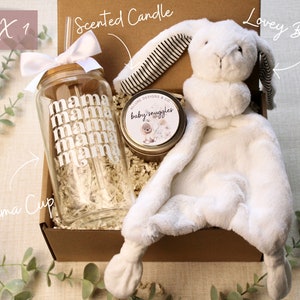 May include: A gift box filled with a white lovey bunny, a scented candle with the text "baby snuggles", and a clear glass cup with the text "mama" repeated multiple times. The box is labeled "BOX 1" and the items are labeled "Lovey Bunny", "Scented Candle", and "Mama Cup".