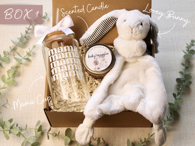 May include: A gift box filled with a white lovey bunny, a scented candle with the text "baby snuggles", and a clear glass cup with the text "mama" repeated multiple times. The box is labeled "BOX 1" and the items are labeled "Lovey Bunny", "Scented Candle", and "Mama Cup".