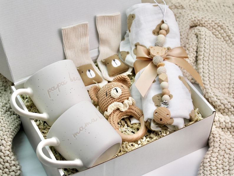 Peut inclure: Une bo&icirc;te cadeau blanche remplie d'un ensemble cadeau de naissance. L'ensemble comprend deux tasses blanches avec les mots "papa bear" et "mama bear" dessus, un hochet en crochet en forme d'ours brun, une paire de chaussettes blanches, une couverture en mousseline blanche avec un imprim&eacute; ours brun et un hochet en bois avec un charme d'ours.