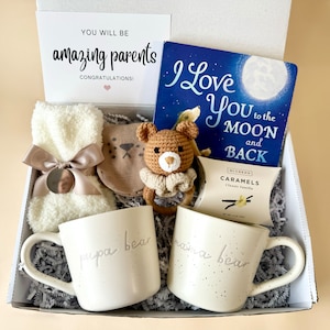 May include: A gift box filled with items for new parents, including two mugs that say "papa bear" and "mama bear", a book titled "I Love You to the Moon and Back", a crocheted teddy bear, a pair of baby booties, and a box of caramels.