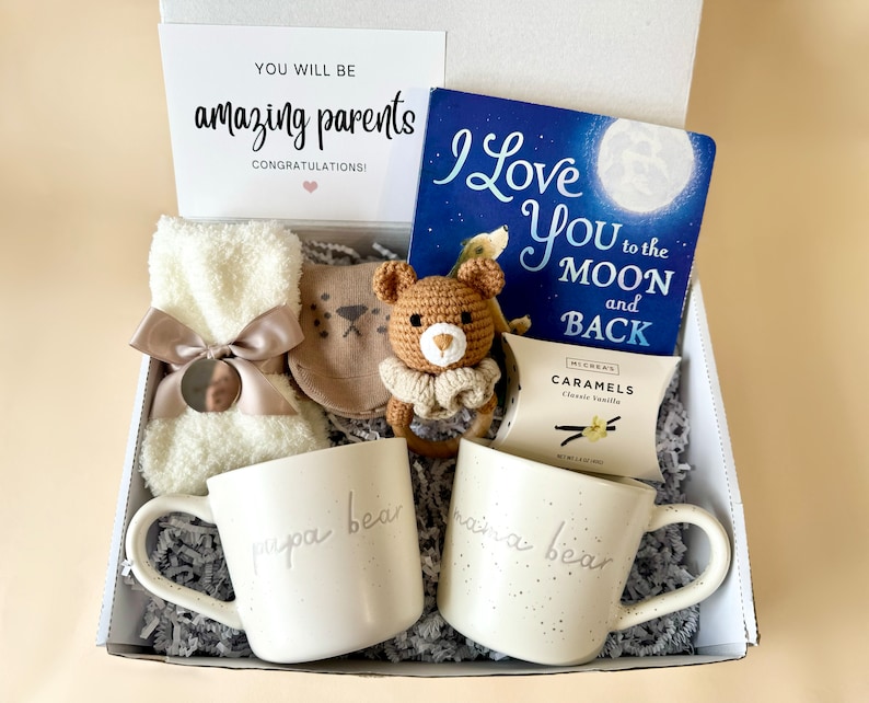 May include: A gift box filled with items for new parents, including two mugs that say "papa bear" and "mama bear", a book titled "I Love You to the Moon and Back", a crocheted teddy bear, a pair of baby booties, and a box of caramels.