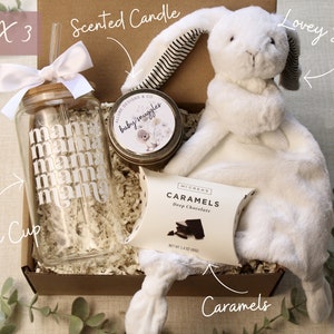 May include: A gift box filled with items for a new mom. The box contains a glass cup with a straw, a white plush bunny, a small jar of scented candle, and a box of chocolates. The cup has the word "mama" printed on it multiple times. The candle jar has the words "baby snuggles" printed on it. The chocolate box has the words "Caramels" and "Deep Chocolate" printed on it.