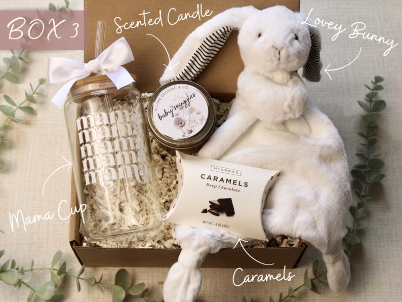May include: A gift box filled with items for a new mom. The box contains a glass cup with a straw, a white plush bunny, a small jar of scented candle, and a box of chocolates. The cup has the word "mama" printed on it multiple times. The candle jar has the words "baby snuggles" printed on it. The chocolate box has the words "Caramels" and "Deep Chocolate" printed on it.
