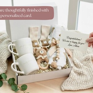 K&ouml;nnte beinhalten: Eine wei&szlig;e Geschenkbox mit Babyartikeln, darunter zwei wei&szlig;e Tassen, Socken, ein geh&auml;keltes B&auml;renrassel und ein Baby-Outfit. Eine Karte lautet: "Herzlichen Gl&uuml;ckwunsch! Wir freuen uns so sehr f&uuml;r Sie! Love, Allure."