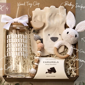 May include: A gift box filled with baby items, including a clear glass water bottle with a straw and the word "mama" repeated on the side, a wooden toy clip with a bird and beads, a pair of beige baby socks, a crocheted bunny rattle, and a box of chocolate caramels.