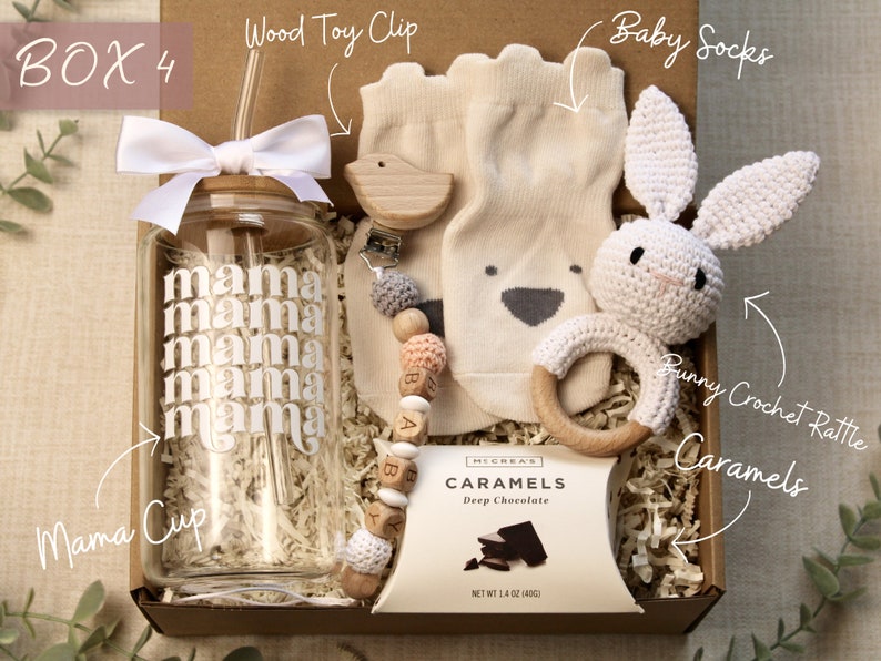 May include: A gift box filled with baby items, including a clear glass water bottle with a straw and the word "mama" repeated on the side, a wooden toy clip with a bird and beads, a pair of beige baby socks, a crocheted bunny rattle, and a box of chocolate caramels.