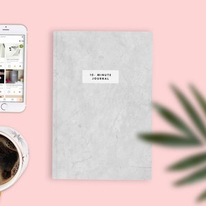 May include: A grey journal with the text "10-Minute Journal" on the front cover. A white smartphone, a cup of coffee, and a green plant are also in the image.