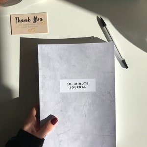 May include: A grey journal with a white label that says "10-Minute Journal". The journal has a textured surface that resembles concrete.