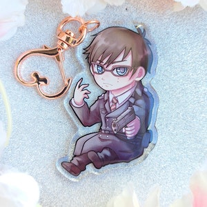 Peut inclure: Un porte-cl&eacute;s en acrylique transparent repr&eacute;sentant un personnage de dessin anim&eacute; portant un costume sombre, des lunettes et tenant une arme &agrave; feu. Le personnage a les cheveux bruns et les yeux bleus.