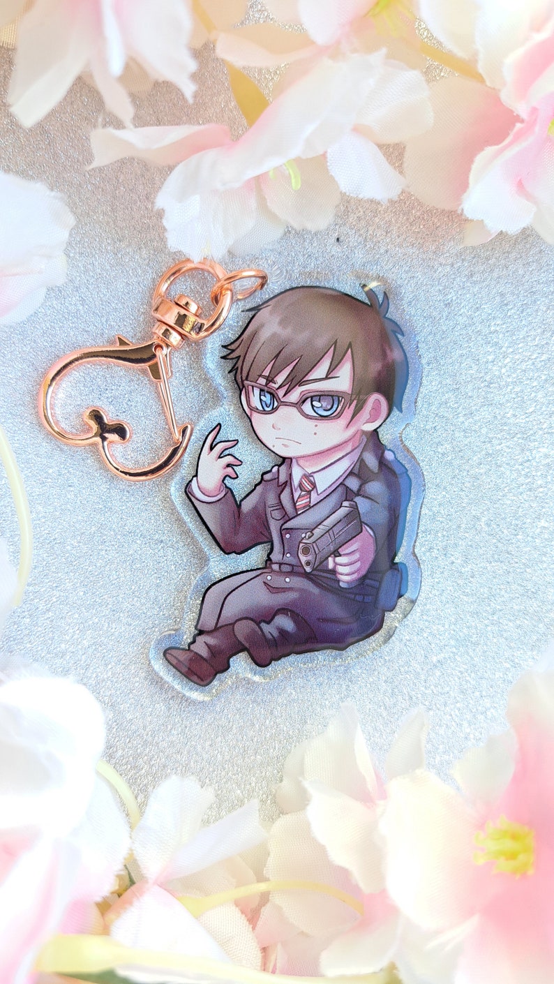 Peut inclure: Un porte-cl&eacute;s en acrylique transparent repr&eacute;sentant un personnage de dessin anim&eacute; portant un costume sombre, des lunettes et tenant une arme &agrave; feu. Le personnage a les cheveux bruns et les yeux bleus.