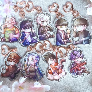 May include: A selection of anime-style character keyrings. Each keyring displays a colourful illustration of a character with a gold-coloured heart-shaped clasp. The keyrings are arranged on a glittery surface, surrounded by pink flowers.
