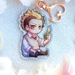 Peut inclure: Un porte-cl&eacute;s repr&eacute;sentant un petit personnage de dessin anim&eacute; avec un fermoir en m&eacute;tal dor&eacute;. Le personnage est un jeune homme aux cheveux blonds, portant une chemise blanche, un gilet en laine marron et une cravate fonc&eacute;e. Il tient un livre vert dans sa main droite.