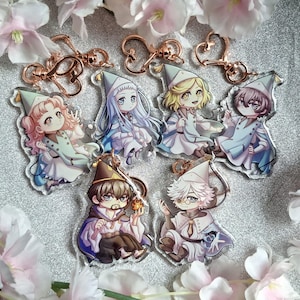 May include: Six acrylic keychains featuring chibi-style anime characters wearing pointed hats and capes. Each charm is attached to a rose gold heart-shaped clasp. The characters have distinct hair colors including pink, blue, blonde, brown, and white.