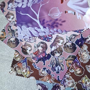 May include: A collection of square microfiber cleaning cloths featuring various anime-style chibi character illustrations. The designs include purple floral patterns, brown backgrounds with multiple figures, and light blue sections with detailed character art.