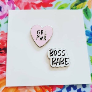 May include: Two enamel pins on a white background. One pin is a pink heart with the text "GRL PWR" in black. The other pin is white with the text "BOSS BABE" in black.