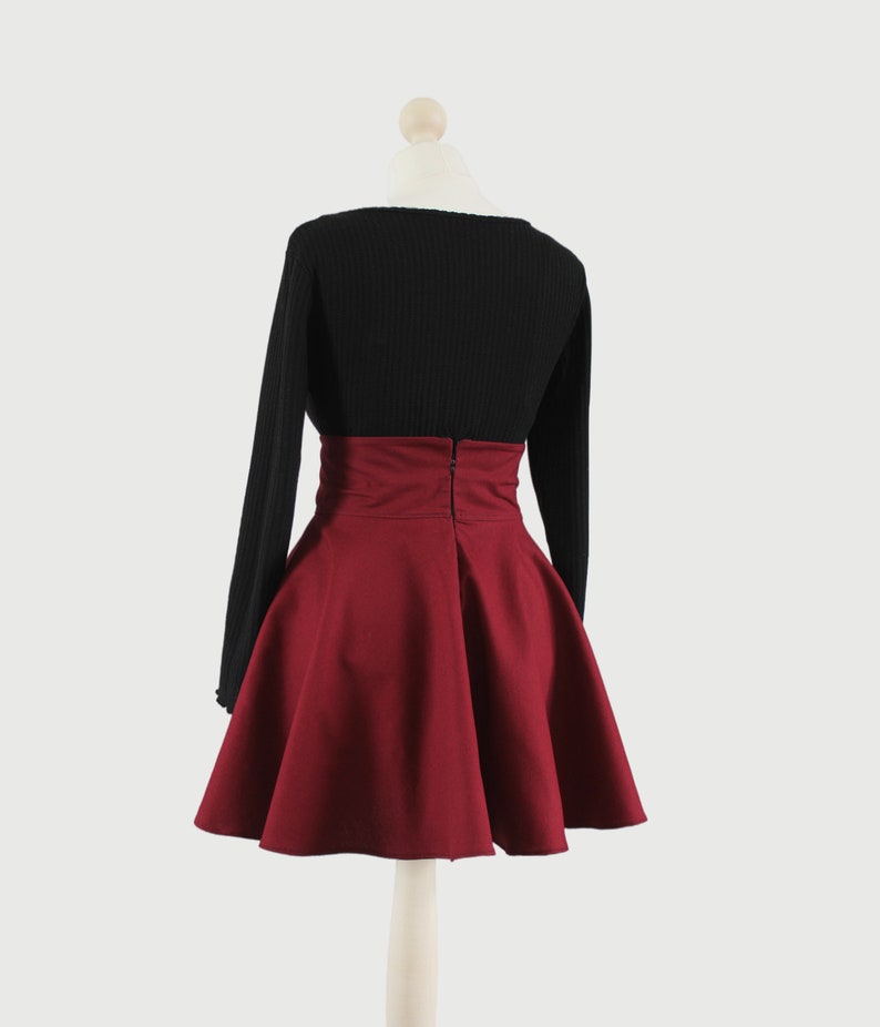 Puede incluir: Una falda de color burdeos de cintura alta con un jersey negro de manga larga. La falda tiene una silueta acampanada y una cremallera en la parte posterior.