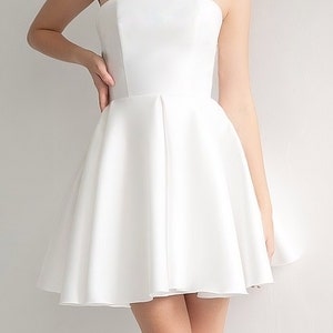 Puede incluir: Un vestido blanco sin tirantes con un corpi&ntilde;o ajustado y una falda completa y fluida. El vestido est&aacute; hecho de una tela lisa y satinada.
