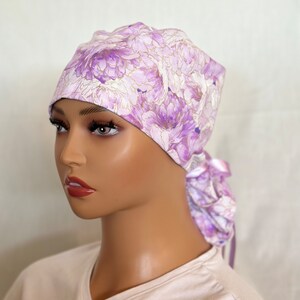 Puede incluir: Un gorro quir&uacute;rgico con estampado floral en tonos lavanda, morado y blanco con detalles dorados. El gorro se ata en la parte posterior con lazos de tela a juego. La tela presenta un patr&oacute;n repetido de flores grandes y estilizadas.