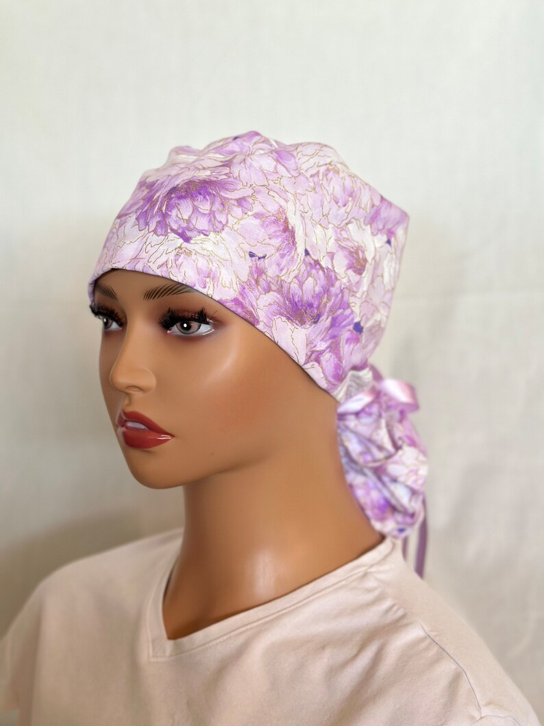 Puede incluir: Un gorro quir&uacute;rgico con estampado floral en tonos lavanda, morado y blanco con detalles dorados. El gorro se ata en la parte posterior con lazos de tela a juego. La tela presenta un patr&oacute;n repetido de flores grandes y estilizadas.