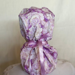 Puede incluir: Un gorro quir&uacute;rgico con un estampado floral en tonos morados y blancos, con detalles dorados. Est&aacute; atado con una cinta lavanda. La tela presenta un dise&ntilde;o de peon&iacute;a. El gorro est&aacute; dise&ntilde;ado para ser usado en la cabeza.