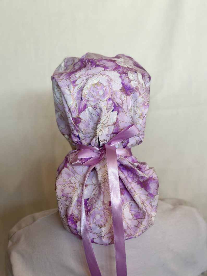 Puede incluir: Un gorro quir&uacute;rgico con un estampado floral en tonos morados y blancos, con detalles dorados. Est&aacute; atado con una cinta lavanda. La tela presenta un dise&ntilde;o de peon&iacute;a. El gorro est&aacute; dise&ntilde;ado para ser usado en la cabeza.