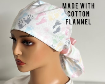 May include: A white cotton flannel scrub cap featuring a pattern of colorful handprints and footprints. The cap is tied at the back with a pink ribbon. Text on the image reads "MADE WITH COTTON FLANNEL".