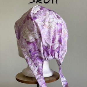 Puede incluir: Un gorro de calavera con estampado floral en tonos morados y blancos con detalles dorados. El gorro se ata en la parte trasera con largas cintas de tela. La palabra "Skull" est&aacute; escrita en negro encima del gorro.