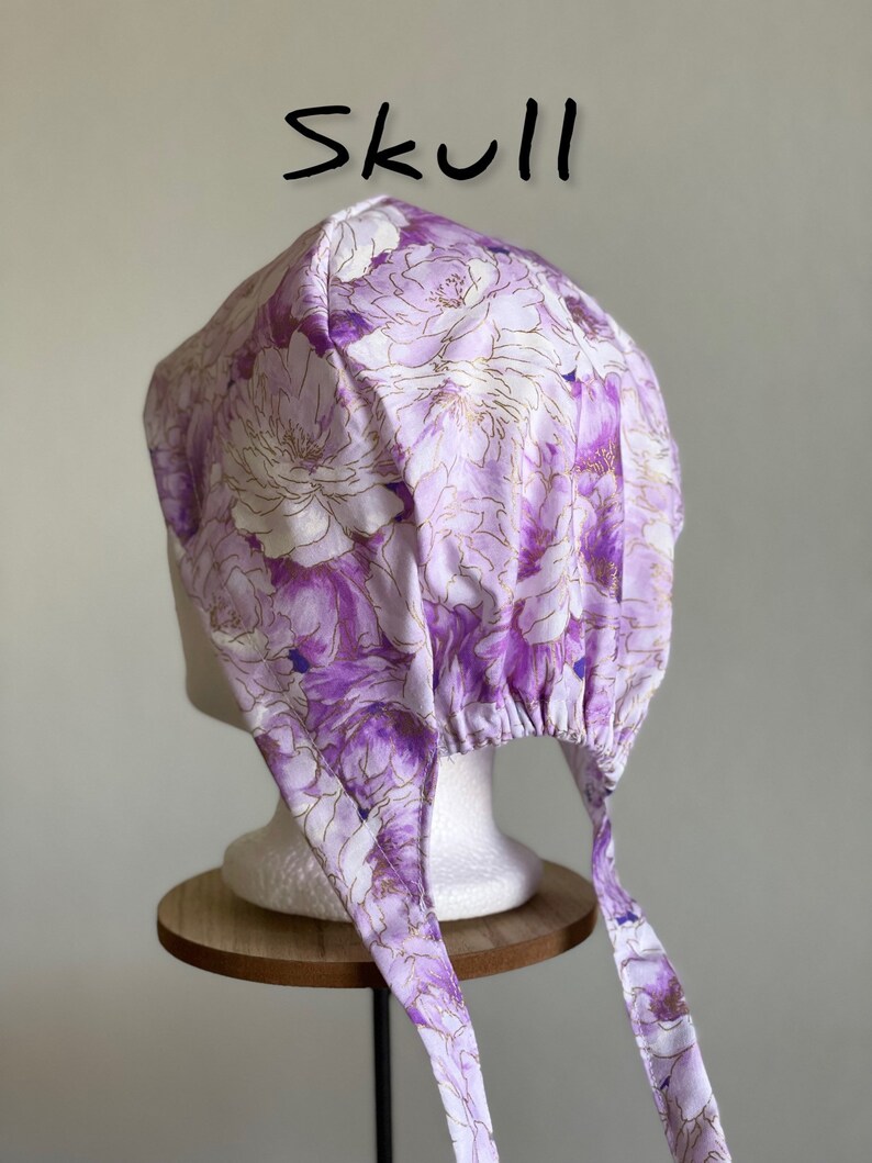 Puede incluir: Un gorro de calavera con estampado floral en tonos morados y blancos con detalles dorados. El gorro se ata en la parte trasera con largas cintas de tela. La palabra "Skull" est&aacute; escrita en negro encima del gorro.