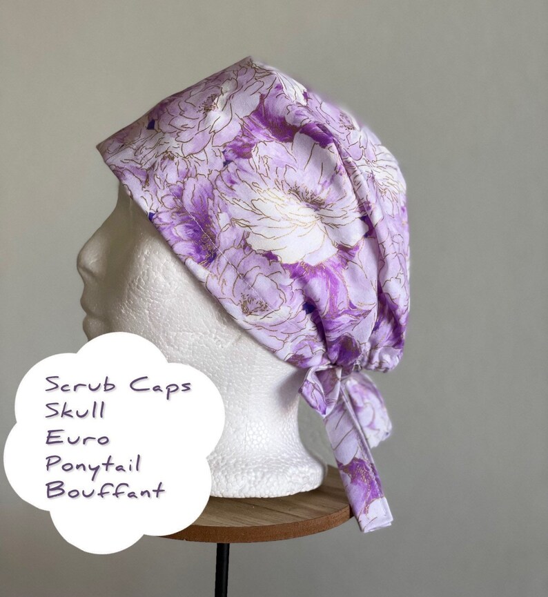 Puede incluir: Un gorro quir&uacute;rgico floral en tonos morados y blancos, con detalles dorados. El gorro est&aacute; atado en la parte trasera y se muestra en una cabeza de maniqu&iacute;. El texto en la imagen dice: "Scrub Caps, Skull, Euro, Ponytail, Bouffant."
