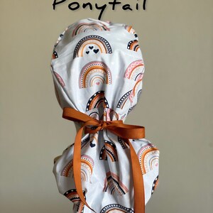 Puede incluir: Un gorro de quir&oacute;fano blanco con cola de caballo con un patr&oacute;n de arco&iacute;ris repetido en tonos naranja, rosa y marr&oacute;n. El gorro est&aacute; atado con una cinta color &oacute;xido. La palabra "Ponytail" est&aacute; escrita en escritura negra encima.