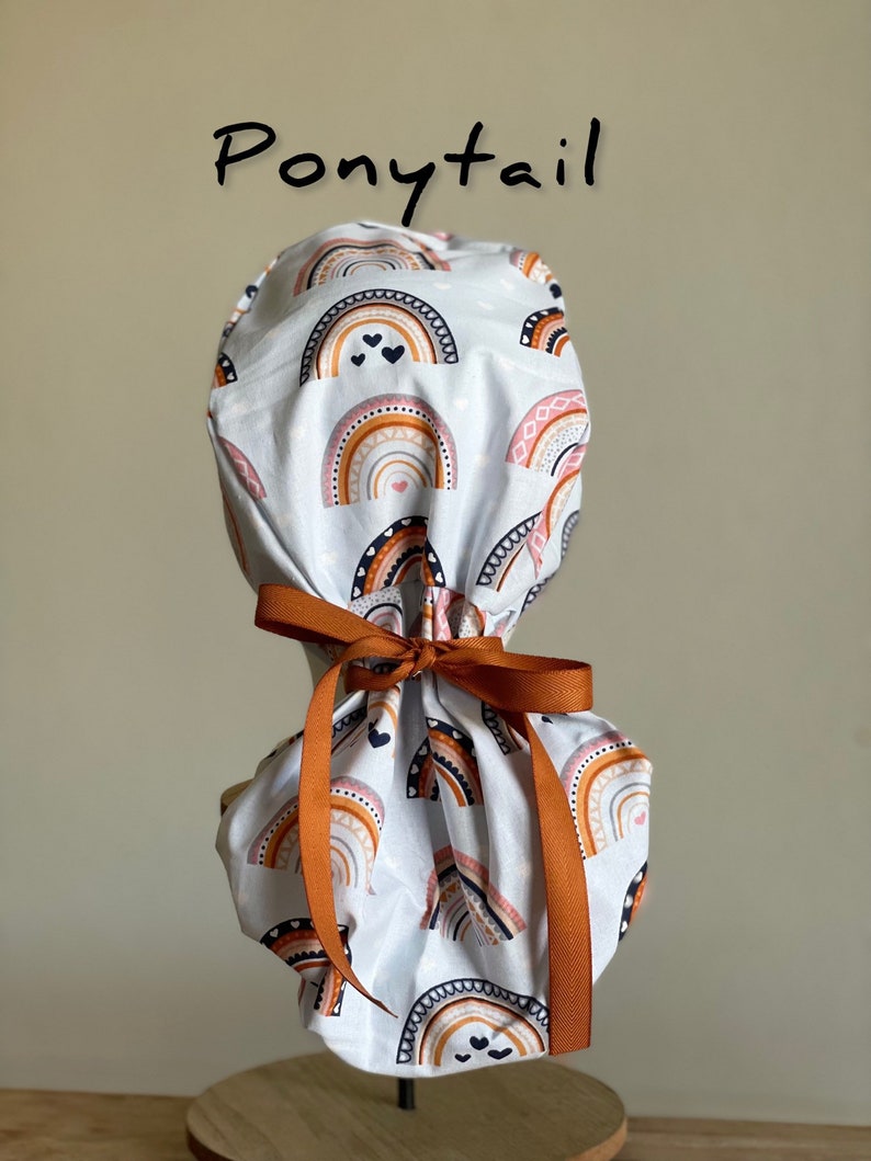 Puede incluir: Un gorro de quir&oacute;fano blanco con cola de caballo con un patr&oacute;n de arco&iacute;ris repetido en tonos naranja, rosa y marr&oacute;n. El gorro est&aacute; atado con una cinta color &oacute;xido. La palabra "Ponytail" est&aacute; escrita en escritura negra encima.