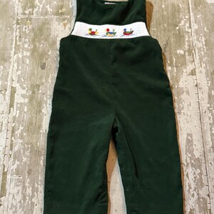 May include: Dark green corduroy baby overalls with a white embroidered band featuring three ducks in boats. The overalls have button closures on the sides and are designed for a young child.