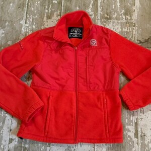 May include: A bright red fleece jacket with a full zipper closure. The jacket has a stand-up collar and two front pockets. The upper chest area has a quilted design. The left chest has a white embroidered "S" logo.