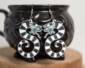 May include: A pair of black and white striped earrings with a green and red creature head on each earring. The earrings are hanging from a black cauldron.