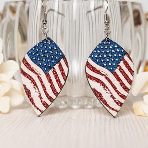 Puede incluir: Un par de pendientes colgantes con un diseño de bandera estadounidense desgastado sobre un fondo blanco. Los pendientes están hechos de cuero y tienen ganchos plateados.