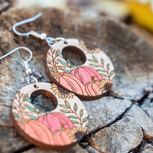 Puede incluir: Un par de pendientes de madera con tem&aacute;tica oto&ntilde;al. Los pendientes son redondos y presentan un dise&ntilde;o de calabazas, girasoles y hojas en tonos naranja, rosa, verde y marr&oacute;n. Los pendientes tienen ganchos plateados.