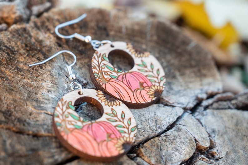 Puede incluir: Un par de pendientes de madera con tem&aacute;tica oto&ntilde;al. Los pendientes son redondos y presentan un dise&ntilde;o de calabazas, girasoles y hojas en tonos naranja, rosa, verde y marr&oacute;n. Los pendientes tienen ganchos plateados.