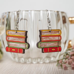 Puede incluir: Un par de pendientes de madera con forma de pila de libros. Los libros están pintados en tonos de rojo, amarillo, verde y marrón. Los pendientes cuelgan de ganchos plateados.