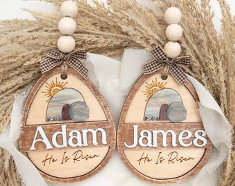 Peut inclure: Deux ornements en forme d'œuf de Pâques en bois avec les noms "Adam" et "James" en lettres blanches. Chaque ornement présente une scène peinte d'un tombeau vide avec un soleil levant et le texte "He Is Risen". Ils sont ornés d'un nœud à carreaux marron et blanc et de perles en bois.