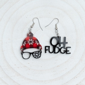 May include: A pair of dangle earrings. One earring features a red and black plaid winter hat with black glasses. The other earring says "OH FUDGE" in black letters. The earrings hang from silver-colored hooks.