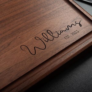 Puede incluir: Tabla de cortar rectangular de madera marrón oscuro. La palabra "Williams" está escrita en cursiva en la superficie, con "EST. 2023" debajo. Una ranura recorre el borde de la tabla. Se ve un cuchillo.