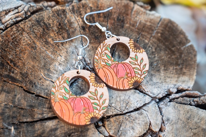 Puede incluir: Un par de pendientes de madera con tem&aacute;tica oto&ntilde;al. Los pendientes son redondos y presentan un dise&ntilde;o de calabazas, girasoles y hojas. Los pendientes est&aacute;n pintados en tonos de naranja, amarillo y verde.