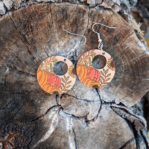 Puede incluir: Un par de pendientes de madera con tem&aacute;tica oto&ntilde;al. Los pendientes son redondos y tienen un recorte en el centro. Est&aacute;n decorados con un patr&oacute;n de calabazas, girasoles y hojas. Los pendientes son de color marr&oacute;n claro con acentos naranja, amarillo y verde.