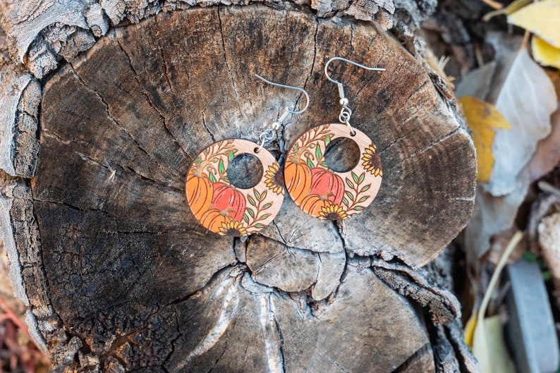 Puede incluir: Un par de pendientes de madera con tem&aacute;tica oto&ntilde;al. Los pendientes son redondos y tienen un recorte en el centro. Est&aacute;n decorados con un patr&oacute;n de calabazas, girasoles y hojas. Los pendientes son de color marr&oacute;n claro con acentos naranja, amarillo y verde.