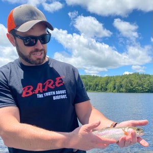 May include: A person wearing a black t-shirt with the text "B.A.R.E. THE TRUTH ABOUT BIGFOOT" in red lettering is holding a rainbow trout fish. The person is standing in front of a lake with a green forest in the background.