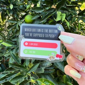 May include: A silver sticker with a green leaf on the top. The sticker has a black background with white text that reads "WHICH BUTTON IS THE BUTTON YOU'RE SUPPOSED TO PUSH?" There are two buttons, one red and one green. The red button has a white X and the text "I AM GROOT." The green button has a white checkmark and the text "THIS ONE." There is a small cartoon drawing of a monster with a speech bubble that says "NO!"