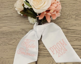 May include: White satin ribbon with pink embroidery that says "Tate & Emma" on one side and "Prom 2024" on the other. The ribbon is tied around a bouquet of pink and white flowers.