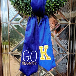 以下が含まれることがあります： 青と金の卒業のタスキに「GO BEARKATS」の文字と反対側に大きな「K」が書かれています。タスキは緑のリースに結ばれています。
