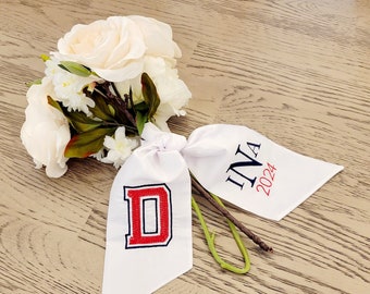 Puede incluir: Un ramo de flores blancas con hojas y tallos verdes, atado con una cinta blanca. La cinta tiene la letra "D" bordada en rojo y negro, y el texto "INA 2024" en negro y rojo.